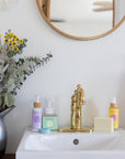 Nourish body lotion, Breathe Everything salve, Rosemary-Mint Everything Soap, Face and Body Cleanser Bar, and Laurentha Aromamist shown together on a white porcelain sink.
