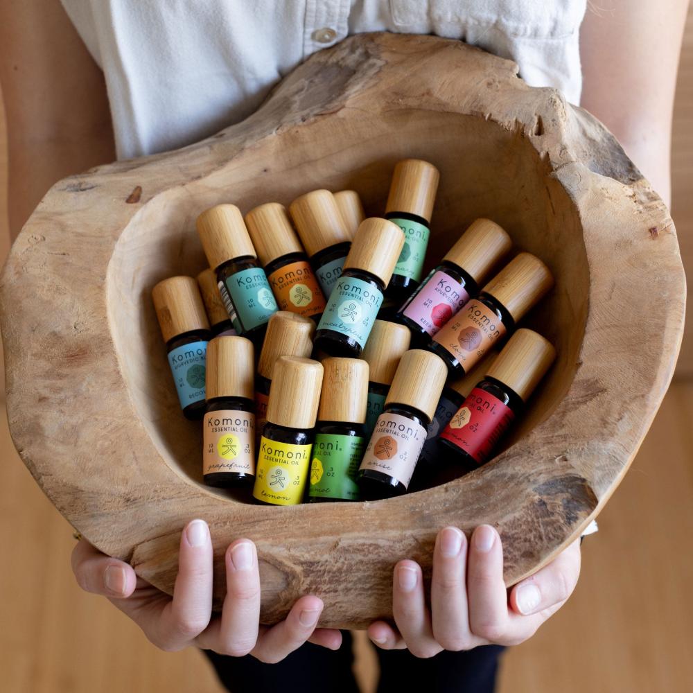 Close up of a woman's hands presenting a rough hewn wooden bowl brimming with colorful 10 mL essential oil vials of every variety featuring elegant bamboo wood tops.