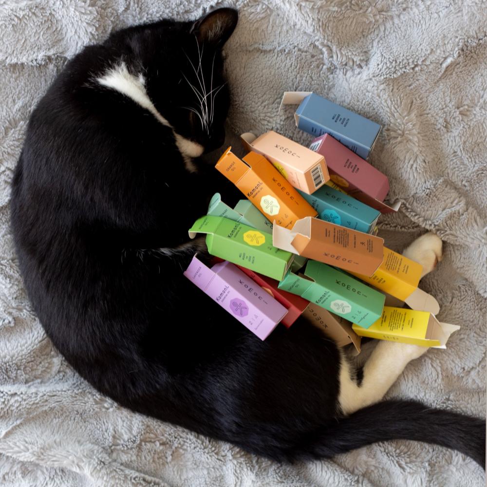 A black cat with white patches shown curled up with all the colorful boxes of Komoni&#39;s pure essential oils.