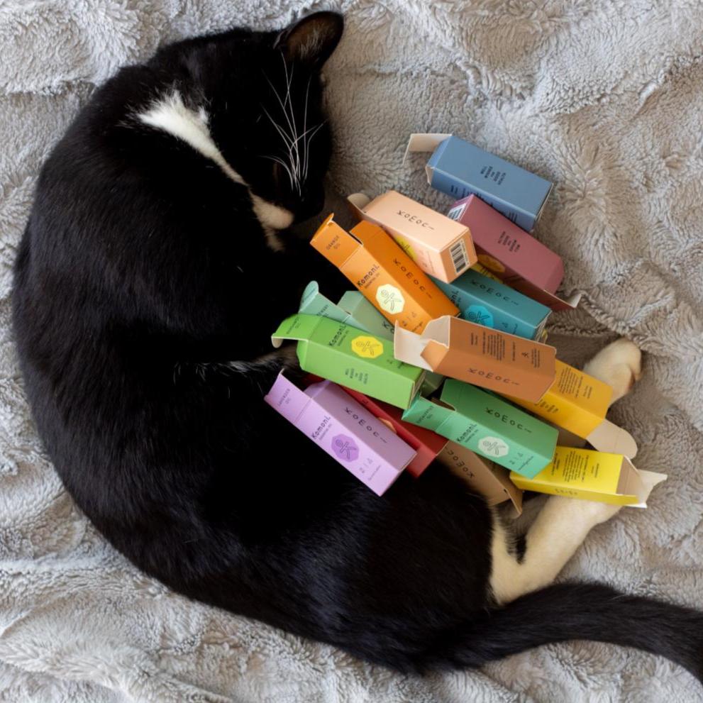 A black cat with white patches shown curled up with all the colorful boxes of Komoni&#39;s pure essential oils.