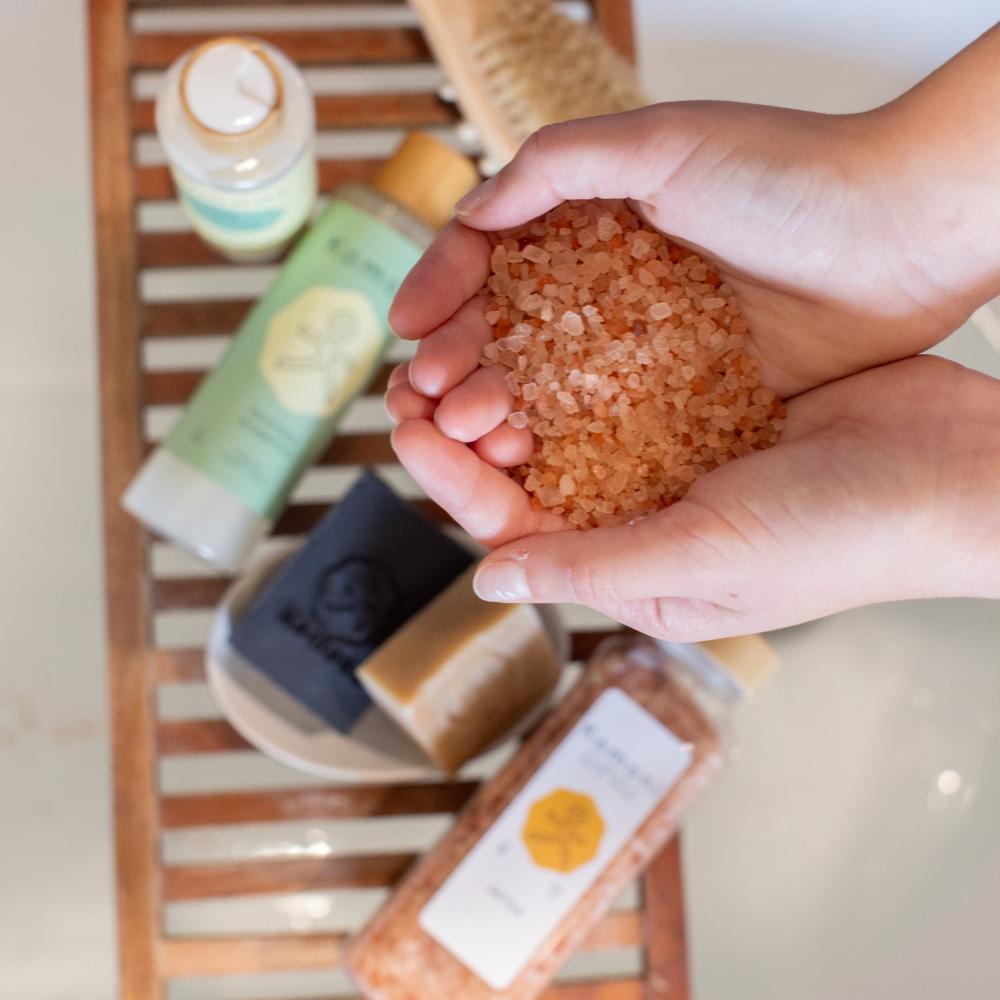 A pair of cupped hands filled with Detox Himalayan Salt Bath Soak with other Komoni bath & body products shown out of focus in the background