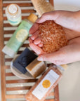 A pair of cupped hands filled with Detox Himalayan Salt Bath Soak with other Komoni bath & body products shown out of focus in the background