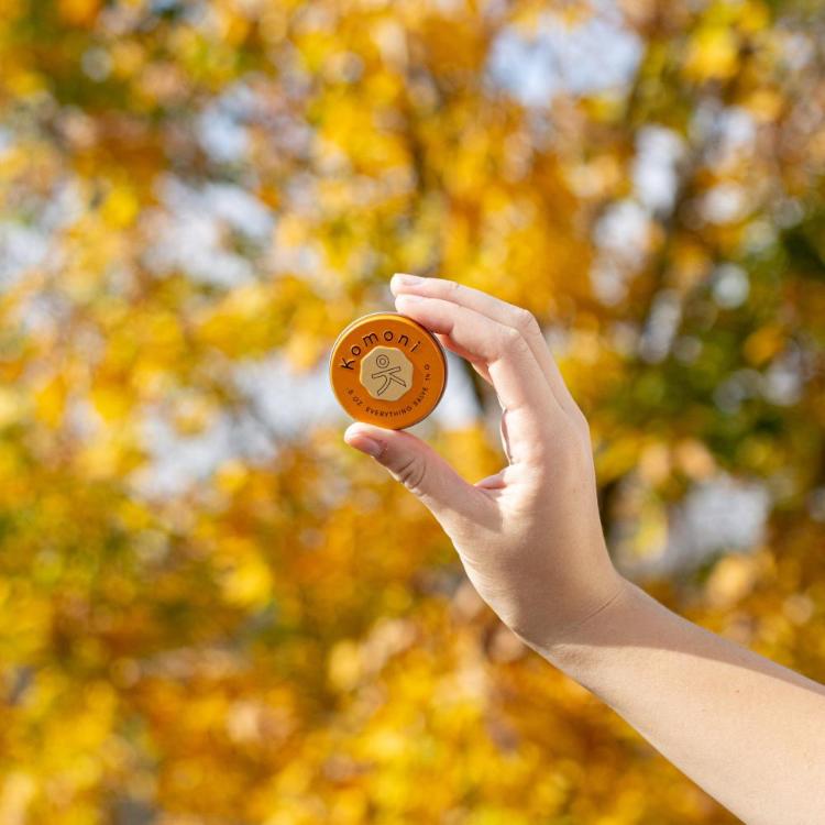 Tin of UPLIFT everything lip and skin pocket-sized salve held up in a hand against a background of matching fall colored leaves.