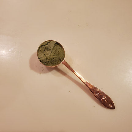 A copper measuring spoon filled with green spinach leaf powder.