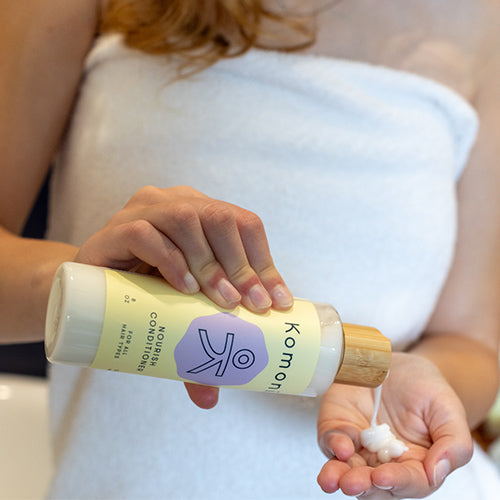 A woman in a bath towel dispensing all-natural nourish shampoo or conditioner with essential oils into her hand from an 8 oz. bottle with elegant bamboo top.