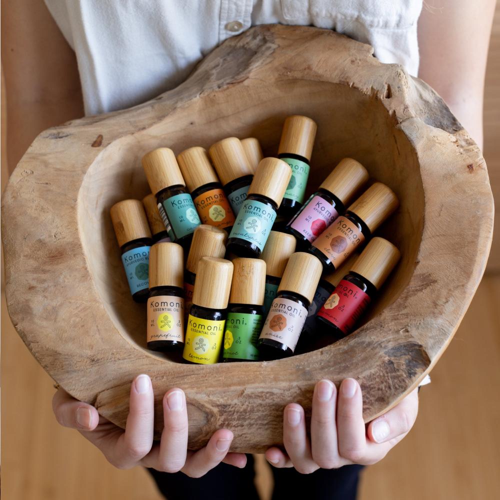 A woman&#39;s hands presenting a rough hewn bowl filled with Komoni&#39;s collection of 100% Pure Essential Oils in colorful 10 mL bottles with beautiful bamboo wood tops.