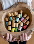 A woman's hands presenting a rough hewn bowl filled with Komoni's collection of 100% Pure Essential Oils in colorful 10 mL bottles with beautiful bamboo wood tops.