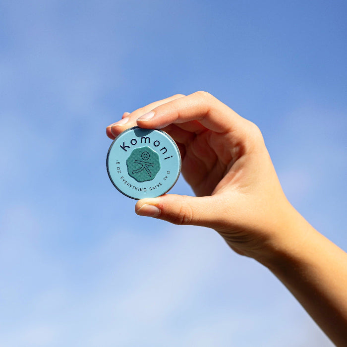 A hand holding tin of BREATHE Everything salve up against a clear blue sky.