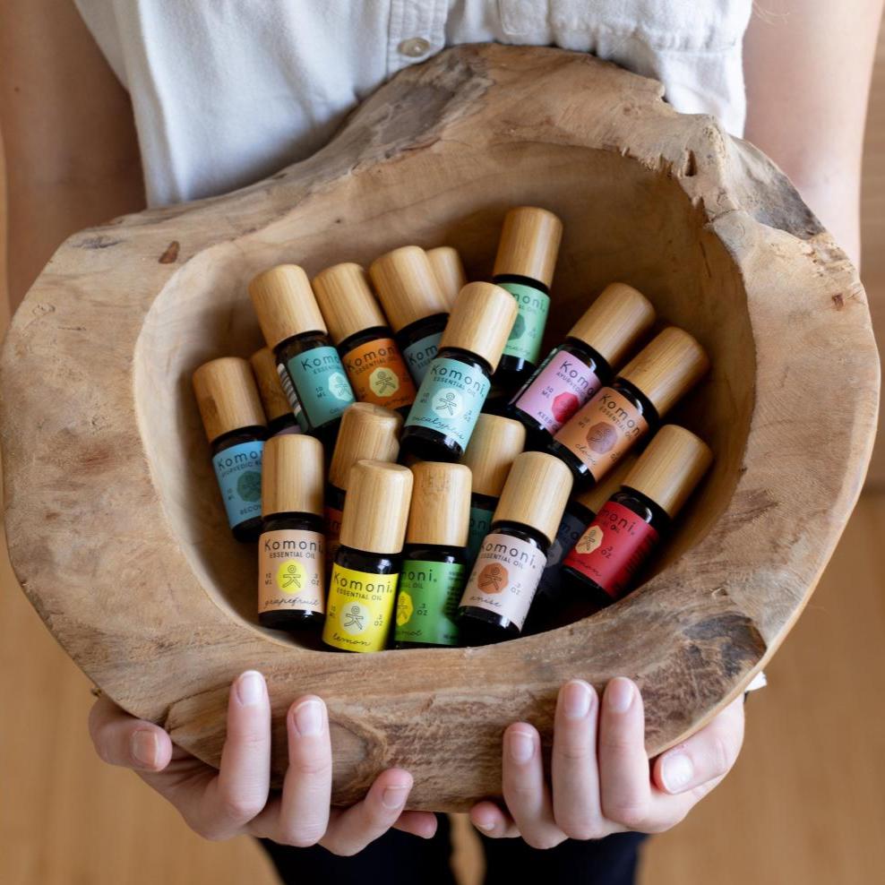 A woman&#39;s hands presenting a rough hewn bowl filled with Komoni&#39;s collection of 100% Pure Essential Oils in colorful 10 mL bottles with beautiful bamboo wood tops.