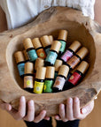 A woman's hands presenting a rough hewn bowl filled with Komoni's collection of 100% Pure Essential Oils in colorful 10 mL bottles with beautiful bamboo wood tops.