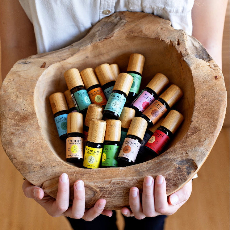 A woman's hands presenting a rough hewn bowl filled with Komoni's collection of 100% Pure Essential Oils in colorful 10 mL bottles with beautiful bamboo wood tops.