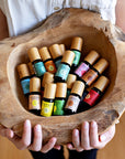 A woman's hands presenting a rough hewn bowl filled with Komoni's collection of 100% Pure Essential Oils in colorful 10 mL bottles with beautiful bamboo wood tops.